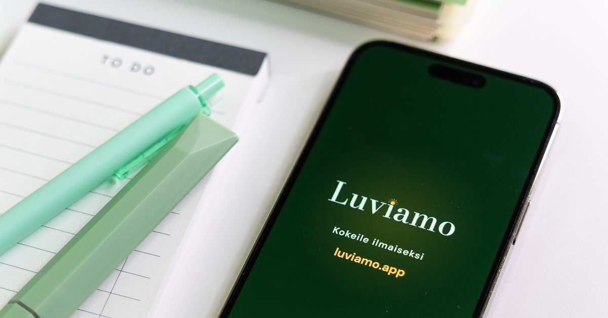 Älypuhelin, jonka näytöllä on Luviamon aloitusnäkymä, muistilehtiön ja kynien vieressä